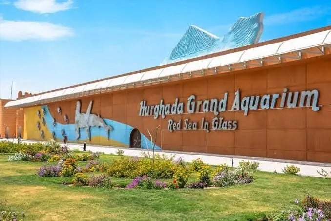 Hurghada Grand Aquarium building with visible signage, surrounded by plants, under a cloudy sky, showcasing marine conservation and family-friendly activities.