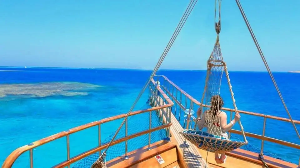 A woman sits relaxed on a hanging swing on the bow of a sailing ship and gazes dreamily out over the endless, glittering sea while the gentle breeze caresses her hair.