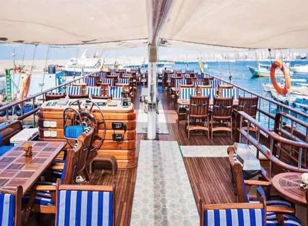 An inviting deck on a sailing boat with several rectangular wooden tables and cozy seating. The white and blue seat cushions lend a maritime atmosphere, while the abundance of wood creates a warm and inviting mood. The harbor stretches out in the background and the sailboat's steering wheel can also be seen, perfectly rounding off the nautical flair.
