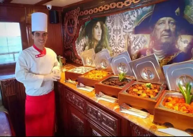 A chef stands proudly in front of an inviting buffet with delicious dishes. In the background, a mural from the movie Pirates of the Caribbean lends the ambience an adventurous pirate atmosphere.