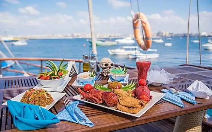 A festively laid table on the deck of a sailing ship, richly plated with fresh seafood specialties. A picturesque harbor stretches out in the background, perfectly rounding off the maritime atmosphere.