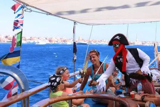 A cheerful family, dressed as pirates, is on the sun-protected deck of a sailing boat with a pirate captain and is clearly enjoying the fun. The silhouette of a town by the sea can be seen in the background, giving the maritime adventure a special backdrop.