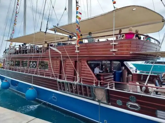The sailing ship is captured from the side as it sails away, while people are on the upper deck and alongside the ship. A small bar with a bartender can be seen at the back, creating an inviting atmosphere.