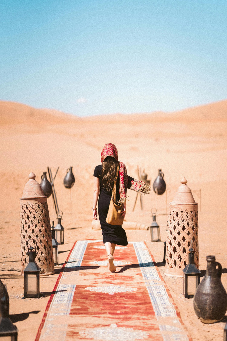 You see a lady from behind in a black desert robe on a red runner in the desert
