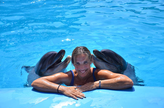 A lady in the pool, 2 dolphins sticking out of the water to the left and right of her, looking cheerful