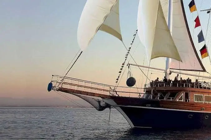 Das Segelschiff liegt ruhig im Meer, während die untergehende Sonne den Himmel in warme Farben taucht. Auf dem Oberdeck genießen zahlreiche Menschen die idyllische Stimmung und den beeindruckenden Sonnenuntergang.