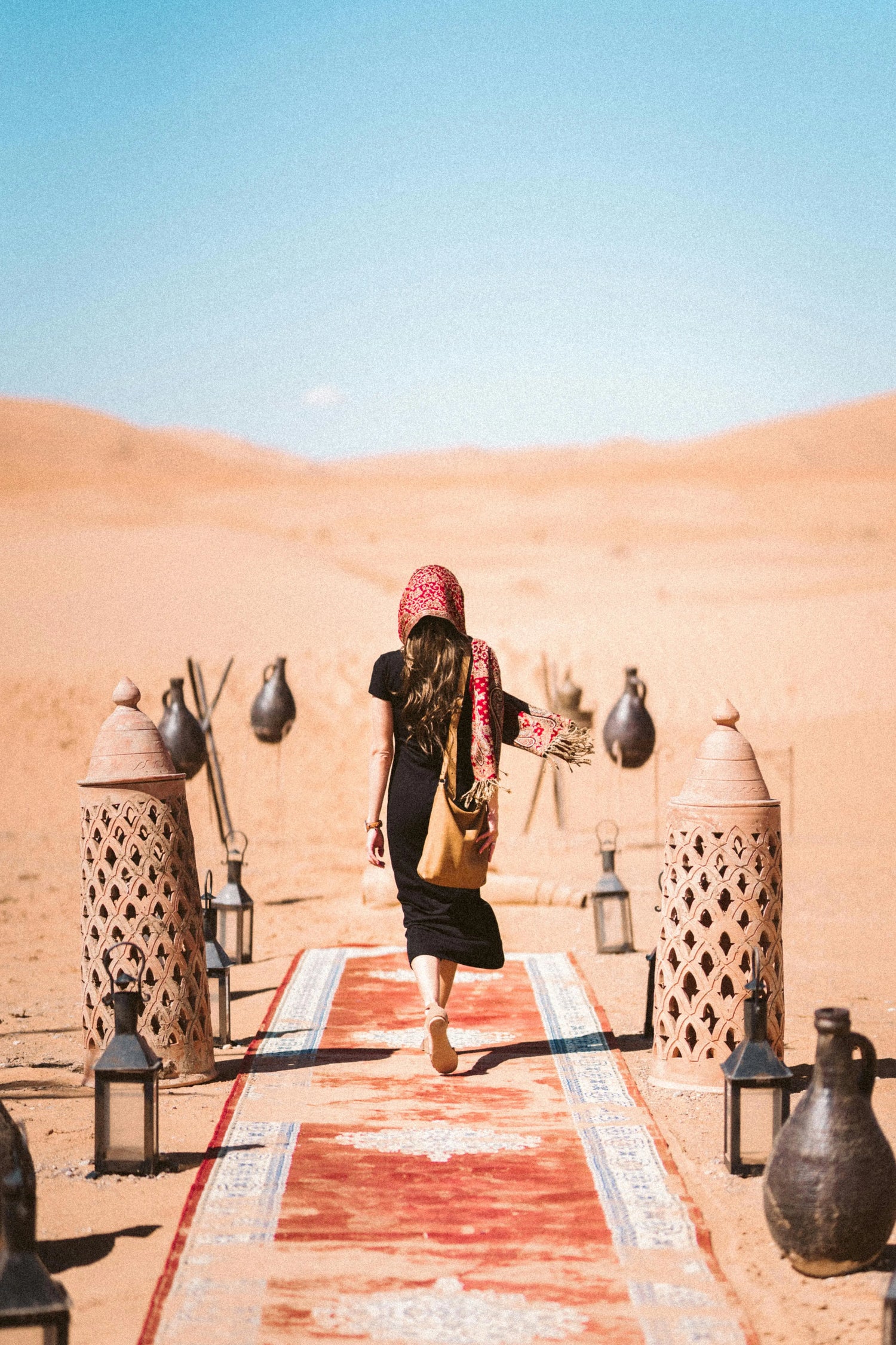 You see a lady from behind in a black desert robe on a red runner in the desert