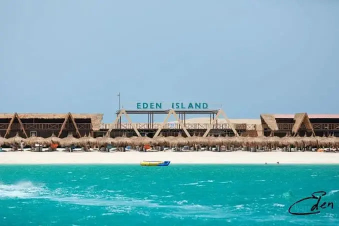 Eden Island Tour: Scenic view of a boat near Hurghada's coastline, with straw umbrellas and a building nearby, highlighting the snorkeling adventure.