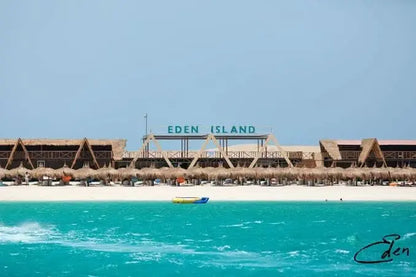 Eden Island Tour: Scenic view of a boat near Hurghada's coastline, with straw umbrellas and a building nearby, highlighting the snorkeling adventure.