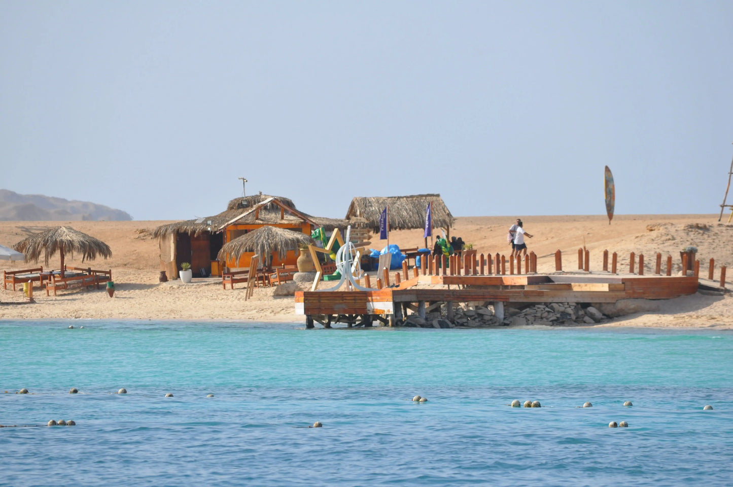 Orange Bay Tour: Image of beach with a dock, huts, people swimming, and a straw umbrella, reflecting the relaxing and adventurous experience in Hurghada.