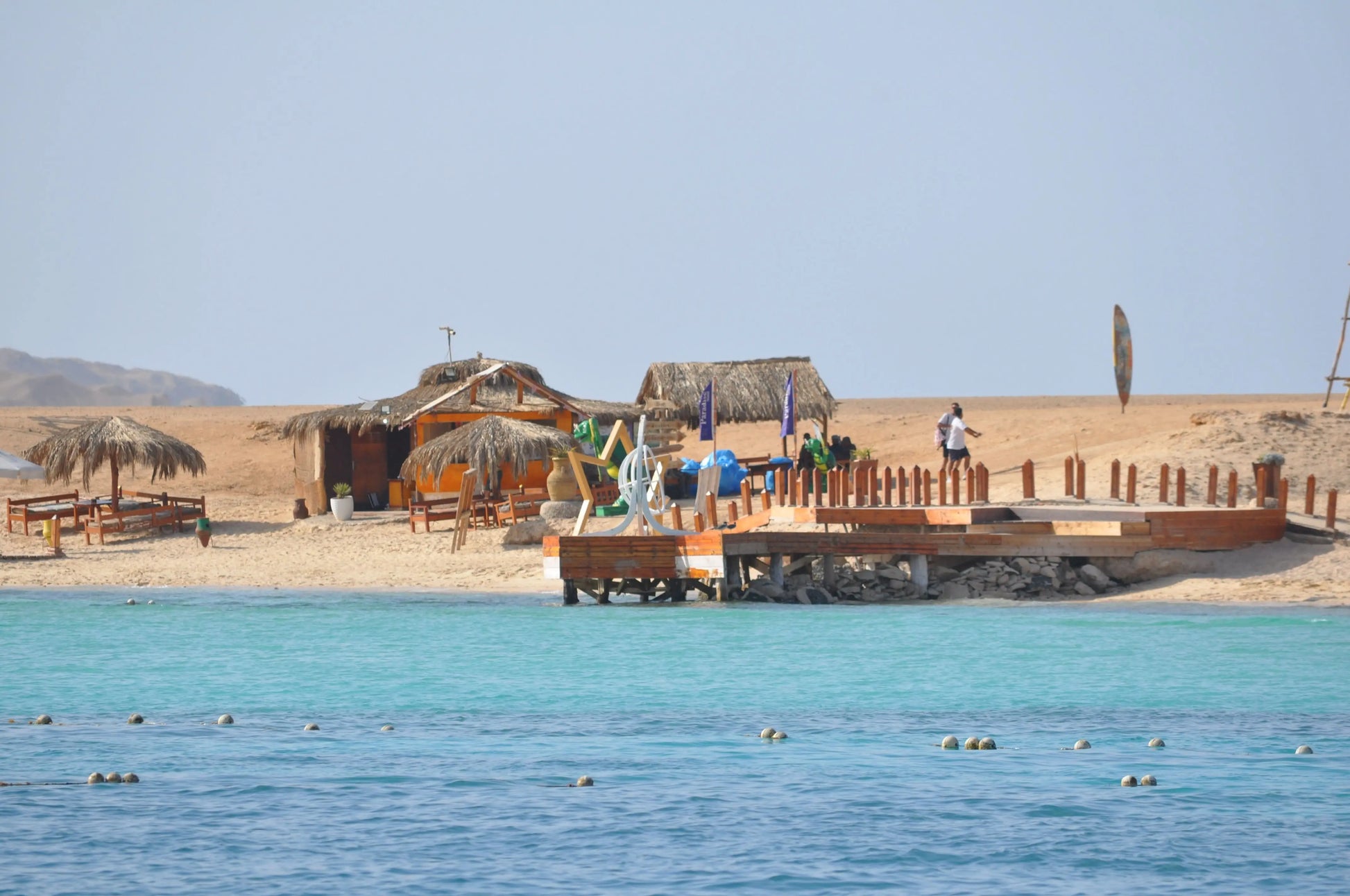 Orange Bay Tour: Image of beach with a dock, huts, people swimming, and a straw umbrella, reflecting the relaxing and adventurous experience in Hurghada.