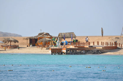 Orange Bay Tour: Image of beach with a dock, huts, people swimming, and a straw umbrella, reflecting the relaxing and adventurous experience in Hurghada.