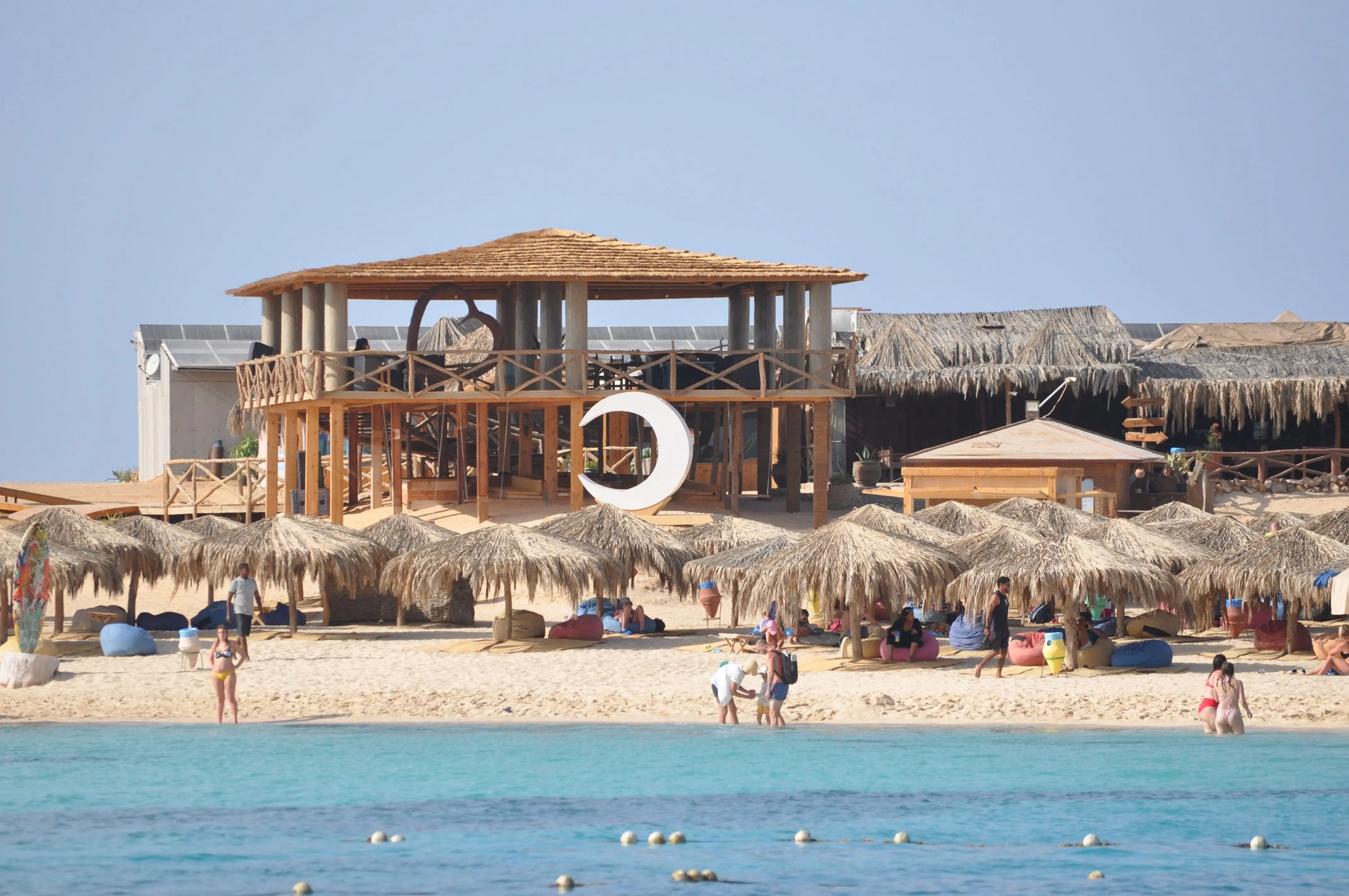 Orange Bay Tour: A serene beach scene with people enjoying the shore, straw umbrellas, and a crescent moon sculpture at a resort in Hurghada.