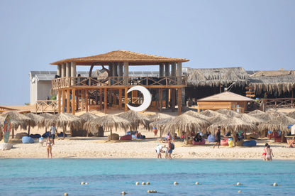 Orange Bay Tour: A serene beach scene with people enjoying the shore, straw umbrellas, and a crescent moon sculpture at a resort in Hurghada.
