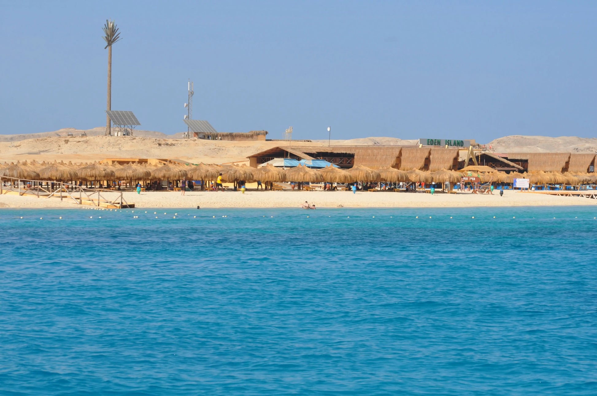 Beach scene with people enjoying Orange Bay Tour in Hurghada; features a beach, building with solar panel, palm tree, and straw umbrellas.