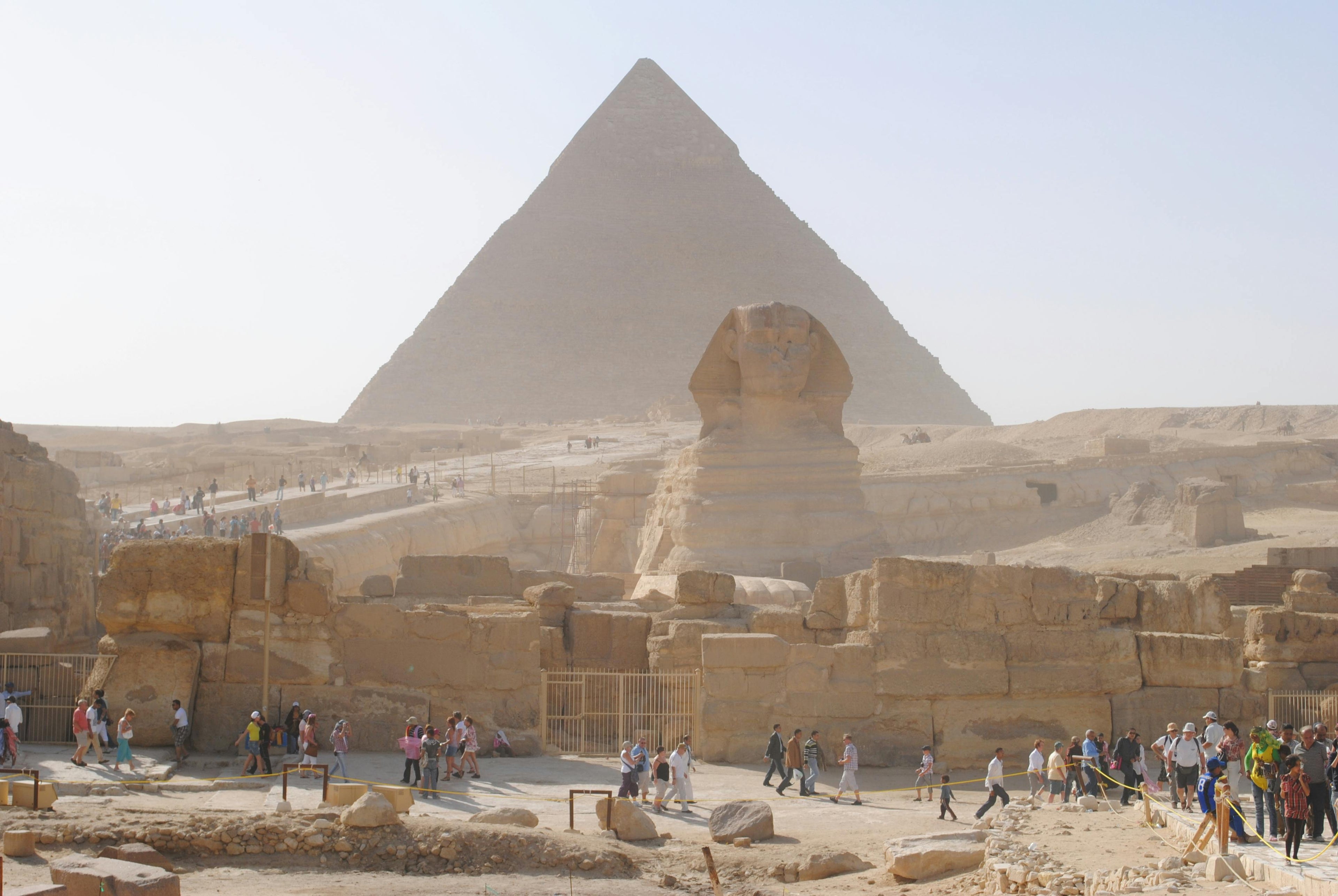A frontal view of the Sphinx and the pyramids in the background. Tourists walk in front of the Sphinx.